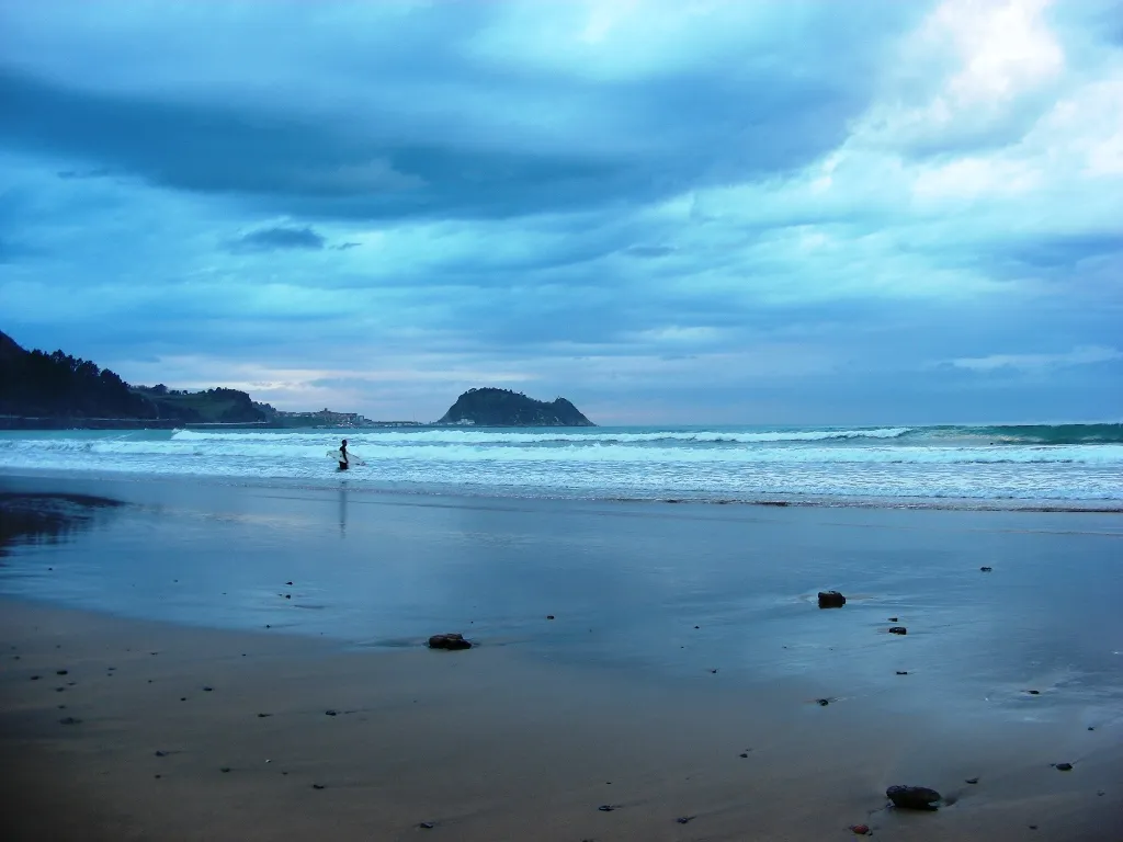 Surfista con la tabla caminando hacia el agua en una playa de Zarautz, bajo un cielo nublado